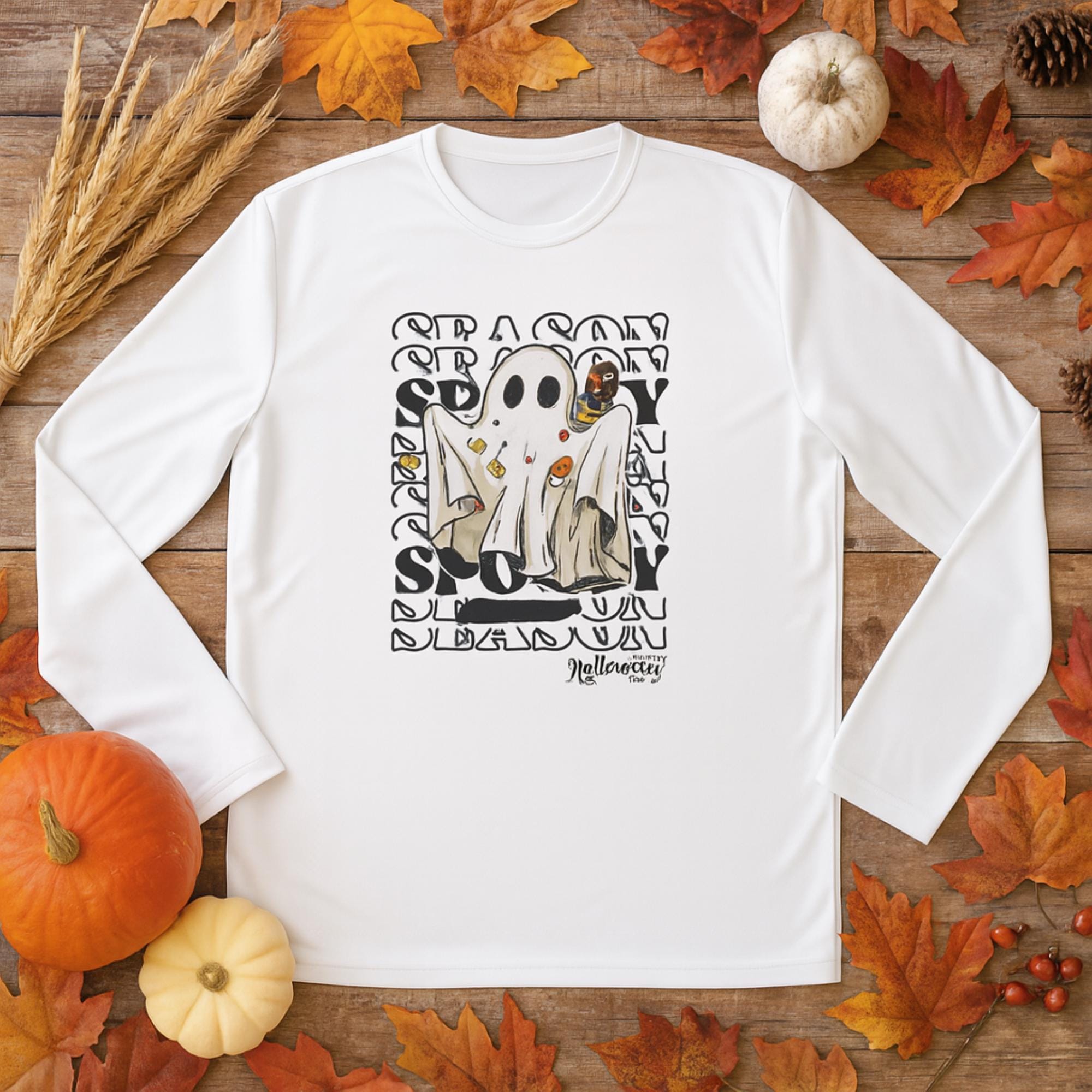 White long-sleeve shirt laid flat on a wooden surface surrounded by autumn leaves, pumpkins, pinecones, and dried wheat stalks. The shirt features a graphic of a ghost costume with candy and Halloween icons, set against a repeating words &#39;SEASON&#39; and &#39;SPOOKY&#39; text background. At the bottom, it says &#39;Halloween&#39; in stylized lettering.