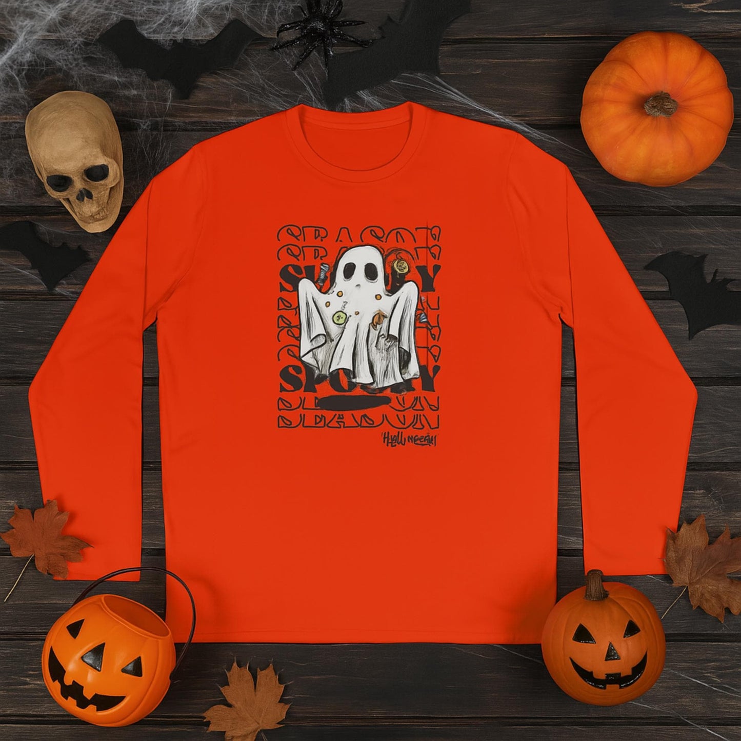 Orange long-sleeve shirt laid flat on a dark wooden surface, featuring a Halloween graphic of a ghost costume with candy, framed by repeating &#39;SEASON SPOOKY&#39; text. The shirt is surrounded by Halloween decorations including jack-o&#39;-lanterns, a skull, black bats, a large spider, cobwebs, and autumn leaves.