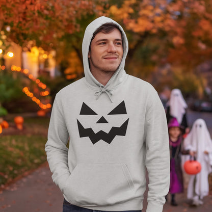Man wearing a light gray hoodie with a black jack-o&#39;-lantern face design, standing outdoors on a neighborhood street decorated with Halloween lights and pumpkins. In the background, children dressed in costumes, including ghosts and a witch, are trick-or-treating among autumn-colored trees.