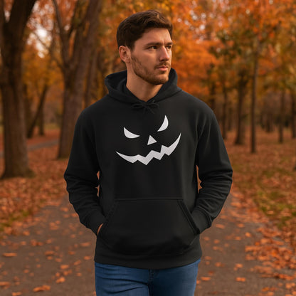 Man walking through a tree-lined path in a park during autumn, wearing a black hoodie with a white jack-o&#39;-lantern face design on the front. The ground is covered with fallen orange leaves, and the trees display vibrant fall foliage.