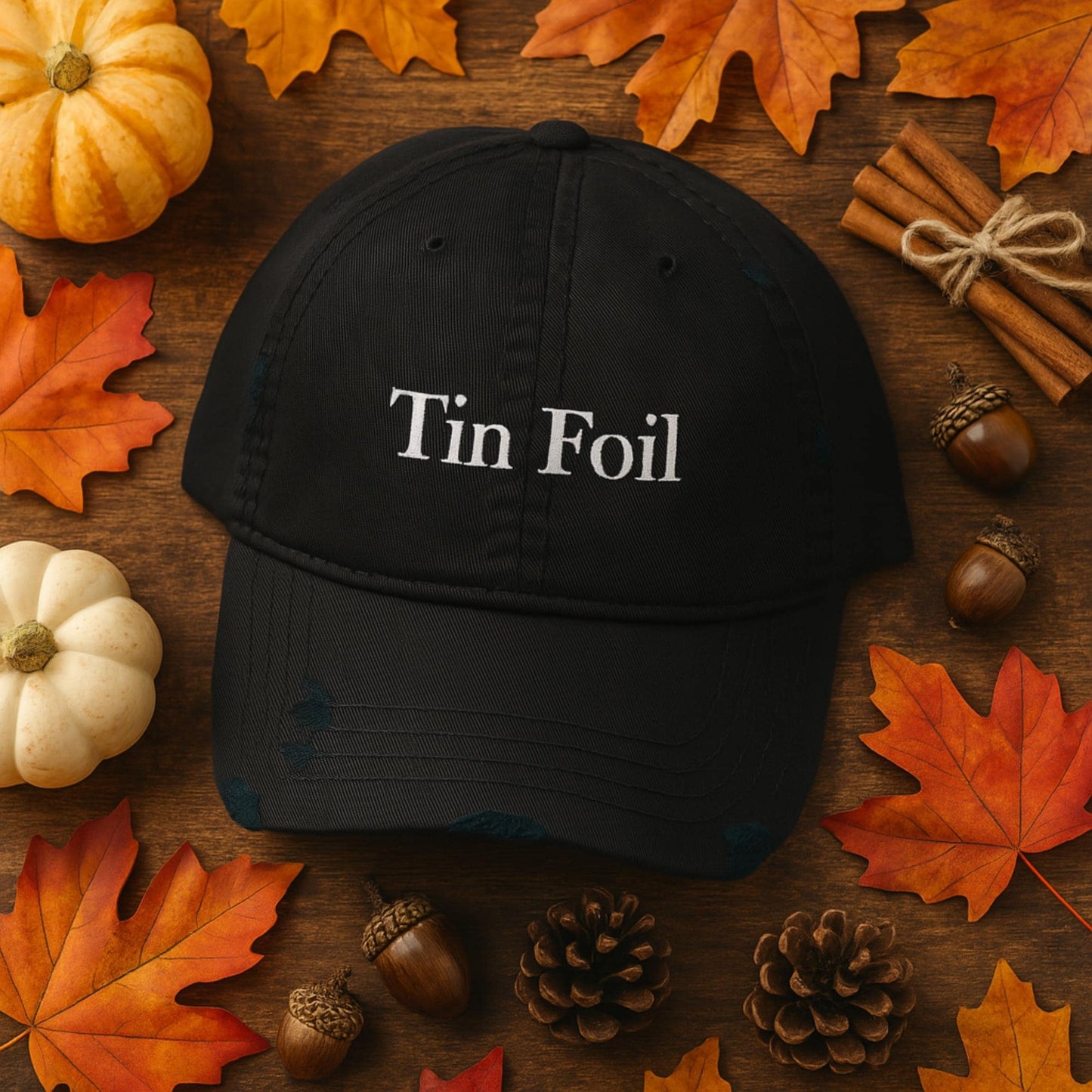 Black baseball cap with the words &#39;Tin Foil&#39; embroidered in white on the front, placed on a wooden surface surrounded by fall-themed decorations including orange and red maple leaves, white and orange pumpkins, cinnamon sticks, acorns, and pinecones.