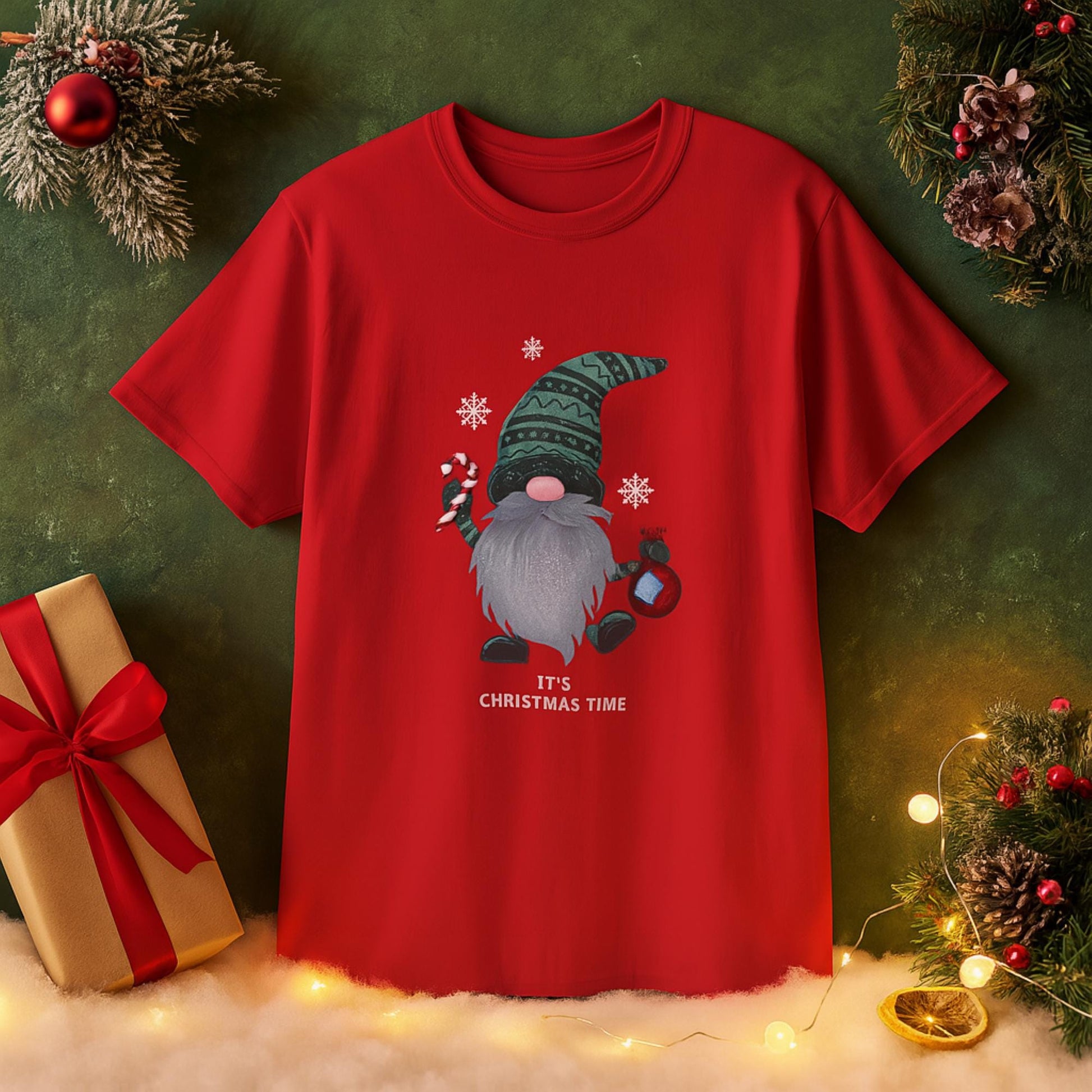 Red T-shirt with a festive Christmas design featuring a gnome with a long gray beard, wearing a green patterned hat, holding a candy cane and a lantern. Snowflakes surround the gnome, and the text &#39;IT&#39;S CHRISTMAS TIME&#39; is printed below. The shirt is displayed against a holiday-themed background with Christmas decorations, gift box, ornaments, lights, and snow.