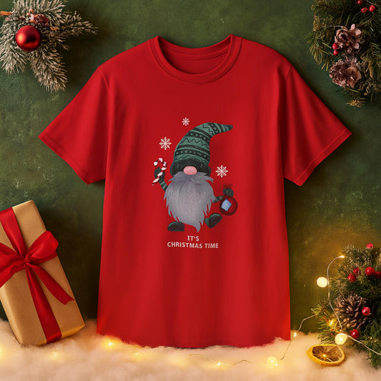 Red T-shirt with a festive Christmas design featuring a gnome with a long gray beard, wearing a green patterned hat, holding a candy cane and a lantern. Snowflakes surround the gnome, and the text &#39;IT&#39;S CHRISTMAS TIME&#39; is printed below. The shirt is displayed against a holiday-themed background with Christmas decorations, gift box, ornaments, lights, and snow.