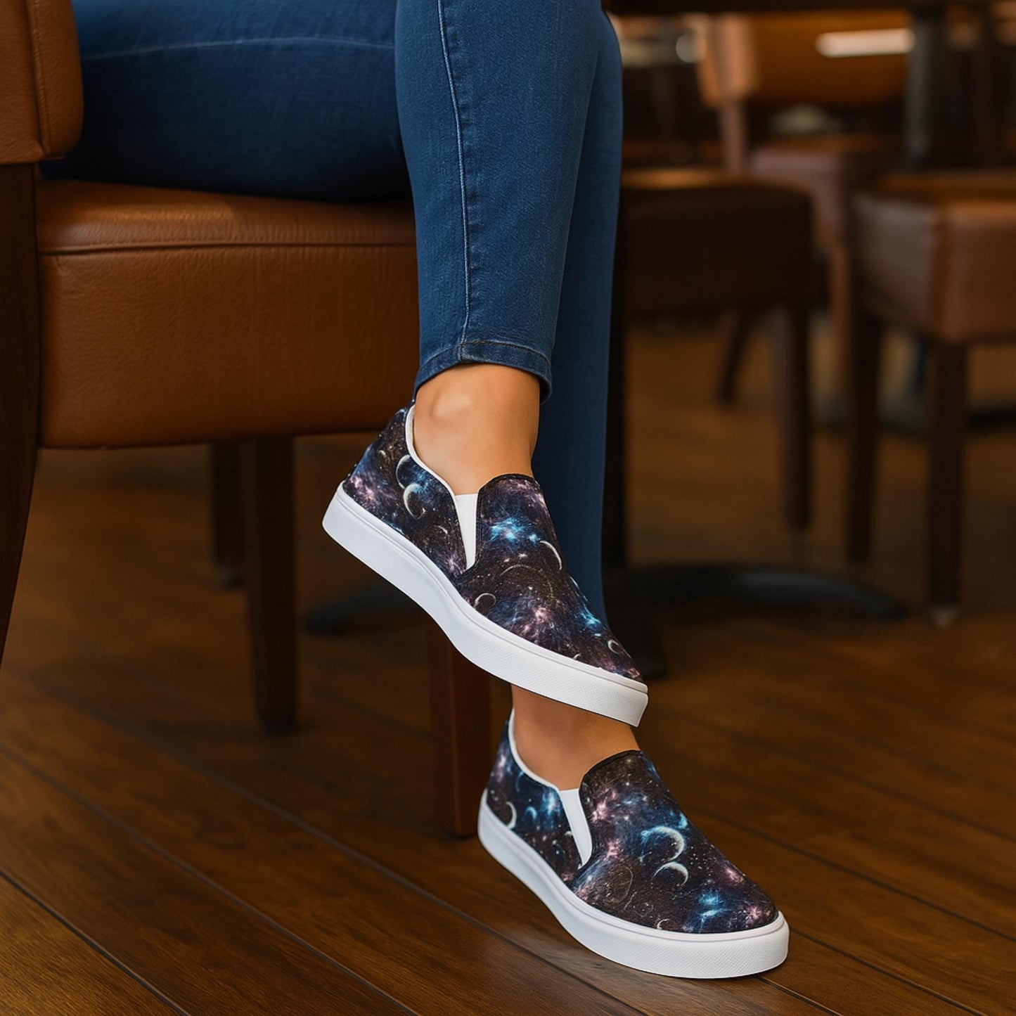 Person seated indoors on a brown leather chair wearing dark blue skinny jeans and galaxy-print slip-on sneakers with white soles. The shoes feature a cosmic design with stars, planets, and nebulae, standing out against the warm-toned wooden floor and furniture.