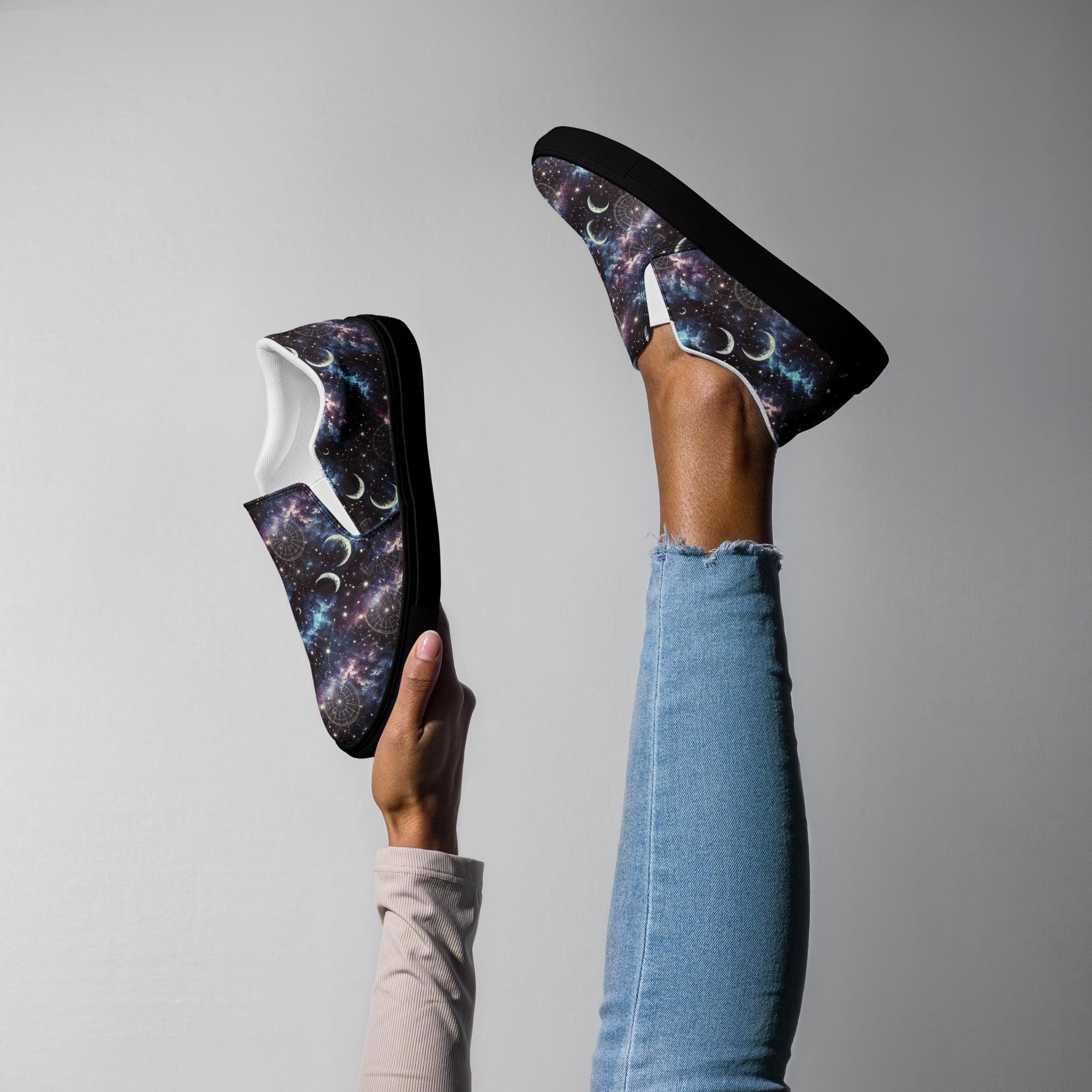 Person playfully posing with one leg raised and holding a matching shoe, both featuring galaxy-themed slip-on sneakers with black soles. The cosmic design displays stars, nebulas, and crescent moons in shades of purple, blue, and white. The person is wearing light blue skinny jeans and a beige long-sleeve top against a plain gray background.