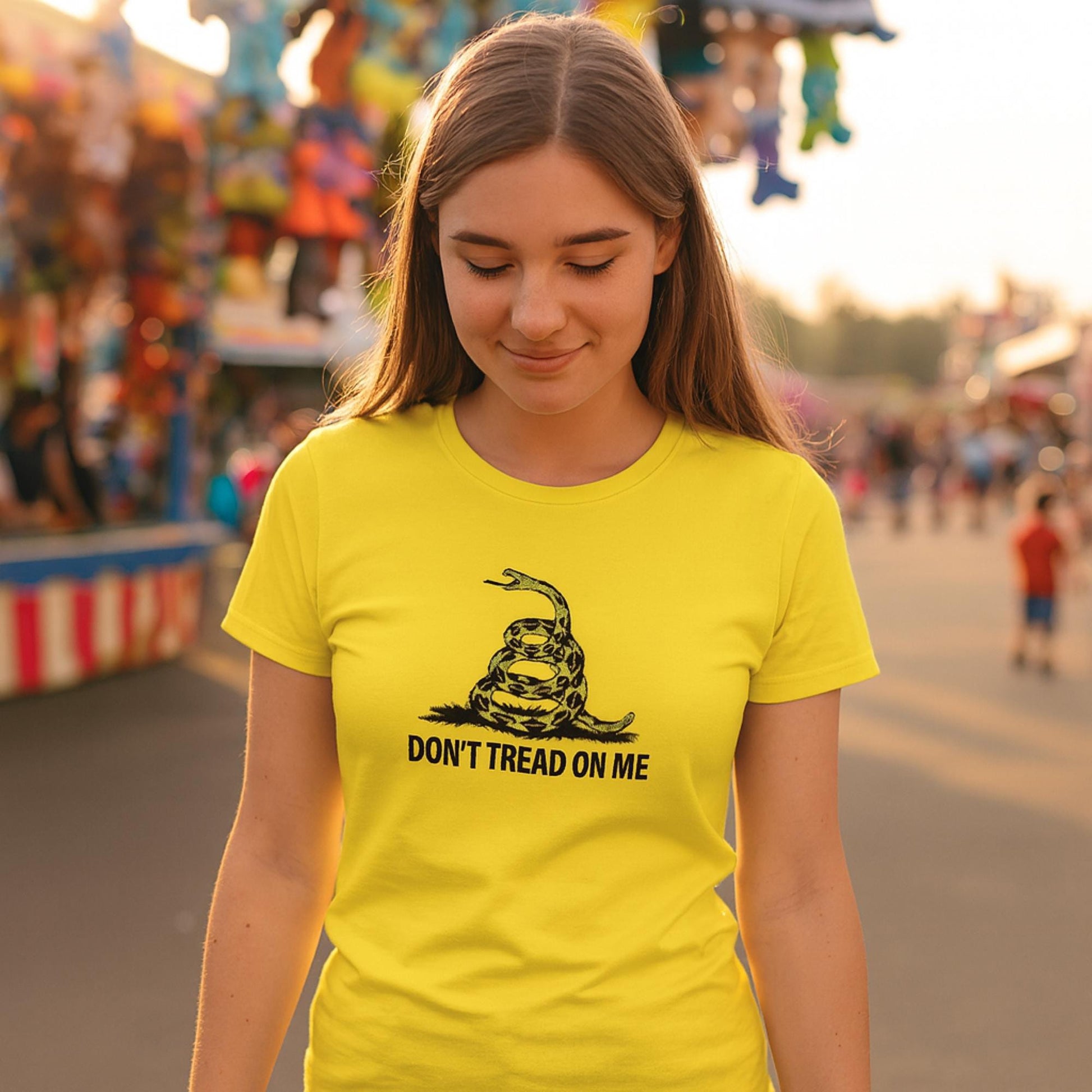 Young woman at a fairground wearing a bright yellow t-shirt with a black graphic of a coiled rattlesnake and the text &quot;DON&#39;T TREAD ON ME&quot; in bold letters. Colorful carnival booths and hanging plush toys fill the background, creating a festive atmosphere.
