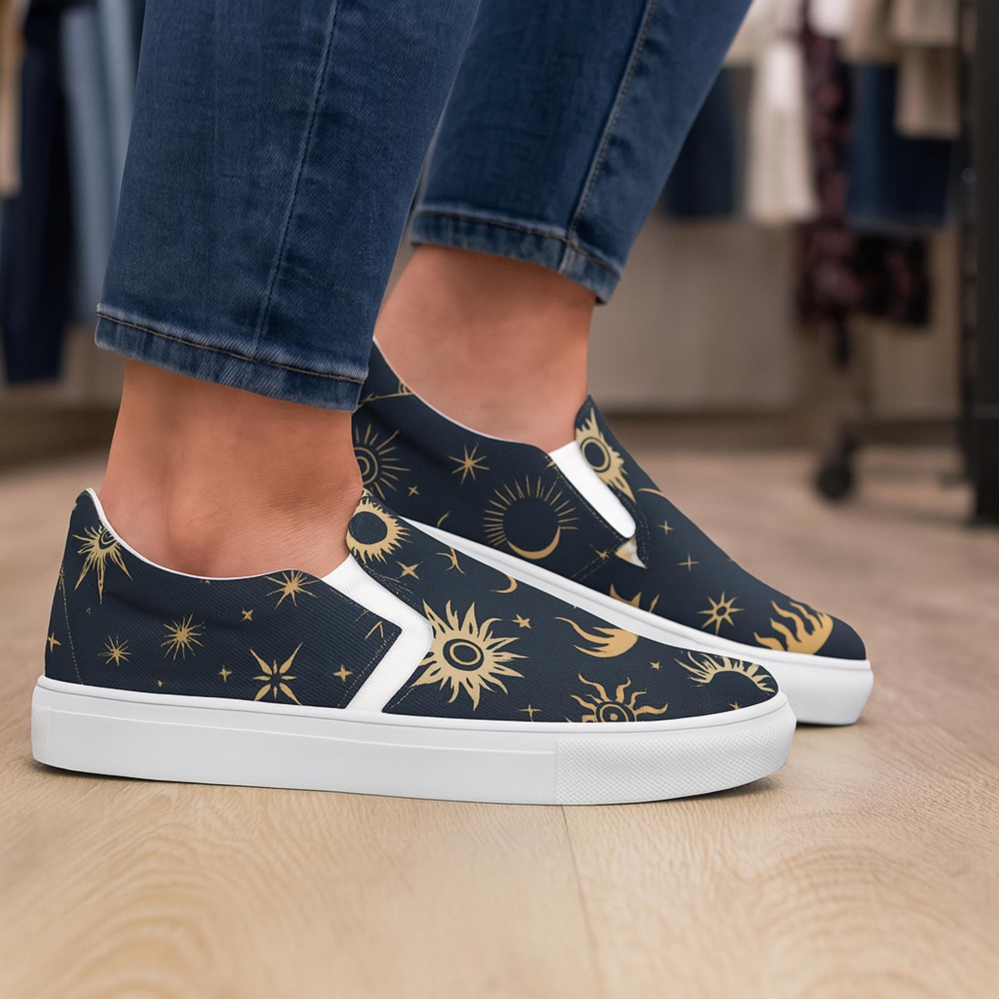 Close-up of a person wearing navy slip-on sneakers with a gold celestial sun and star pattern, paired with cuffed dark blue jeans. The person is standing on a light wood floor in a retail or boutique setting.