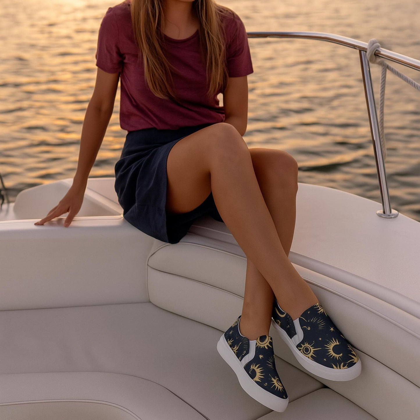 Woman sitting on a white boat seat at sunset, wearing a maroon t-shirt, navy skirt, and navy slip-on shoes featuring a gold celestial pattern with stars and moons. The calm water in the background reflects the warm golden light.