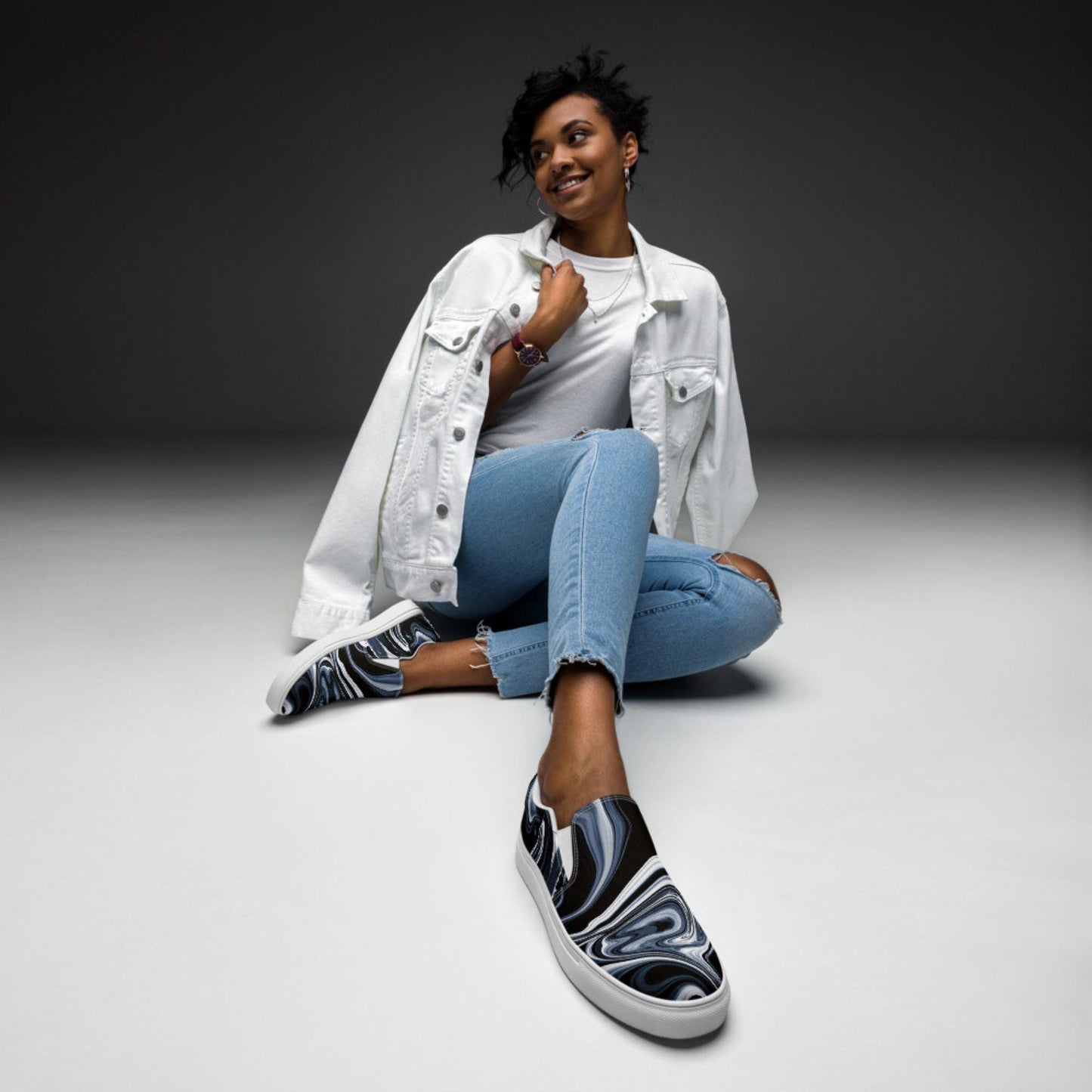 Woman modeling black, white, and gray marbled swirl slip-on canvas sneakers, styled with light blue skinny jeans, a white t-shirt, and a white denim jacket. Fashion-forward casual footwear for women, perfect for street style and everyday wear.