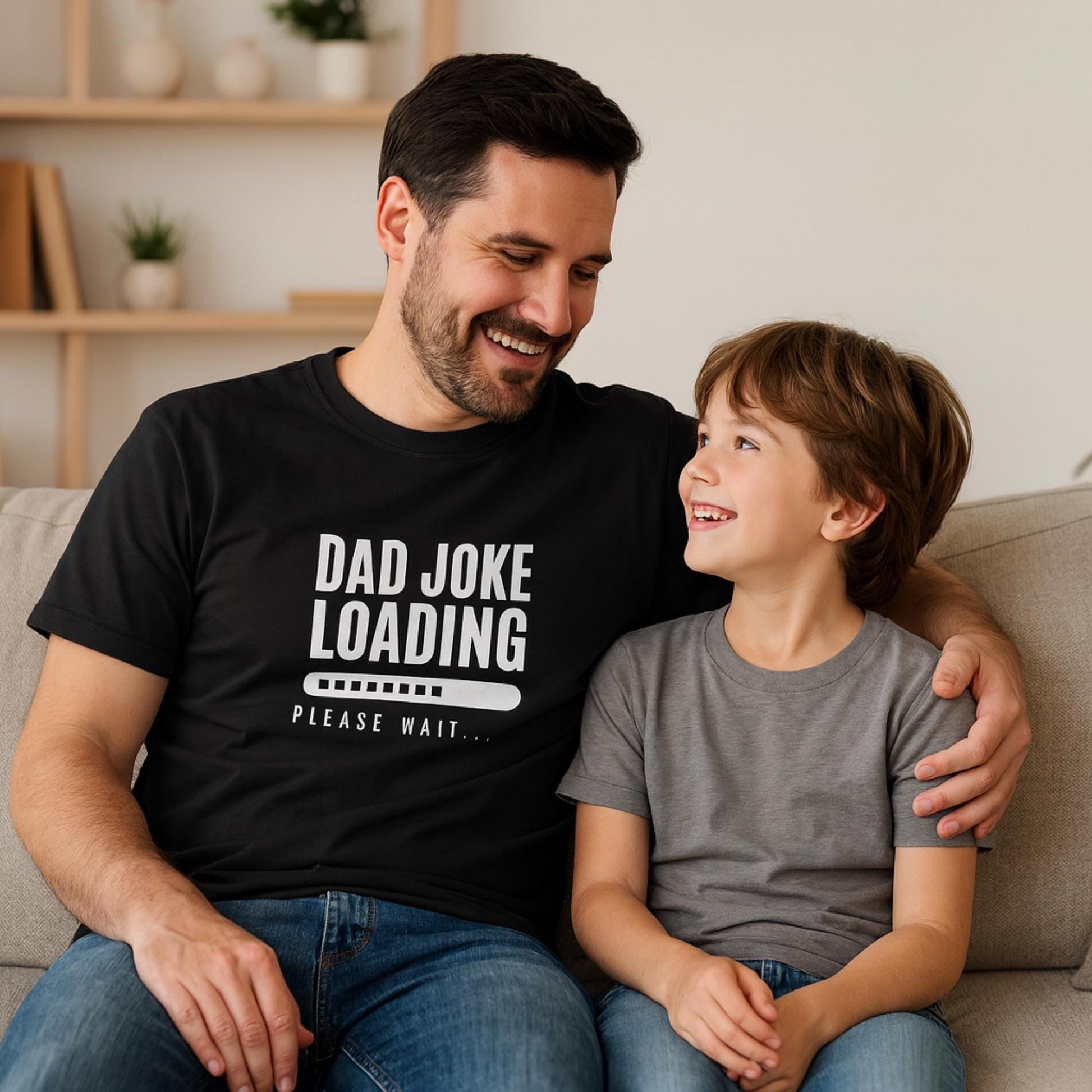 Smiling man wearing a black T-shirt with the text “Dad Joke Loading… Please Wait” and a progress bar graphic in white, sitting on a couch with his arm around a young boy in a gray T-shirt, both looking at each other happily.