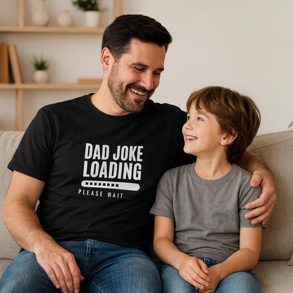 Smiling man wearing a black T-shirt with the text “Dad Joke Loading… Please Wait” and a progress bar graphic in white, sitting on a couch with his arm around a young boy in a gray T-shirt, both looking at each other happily.