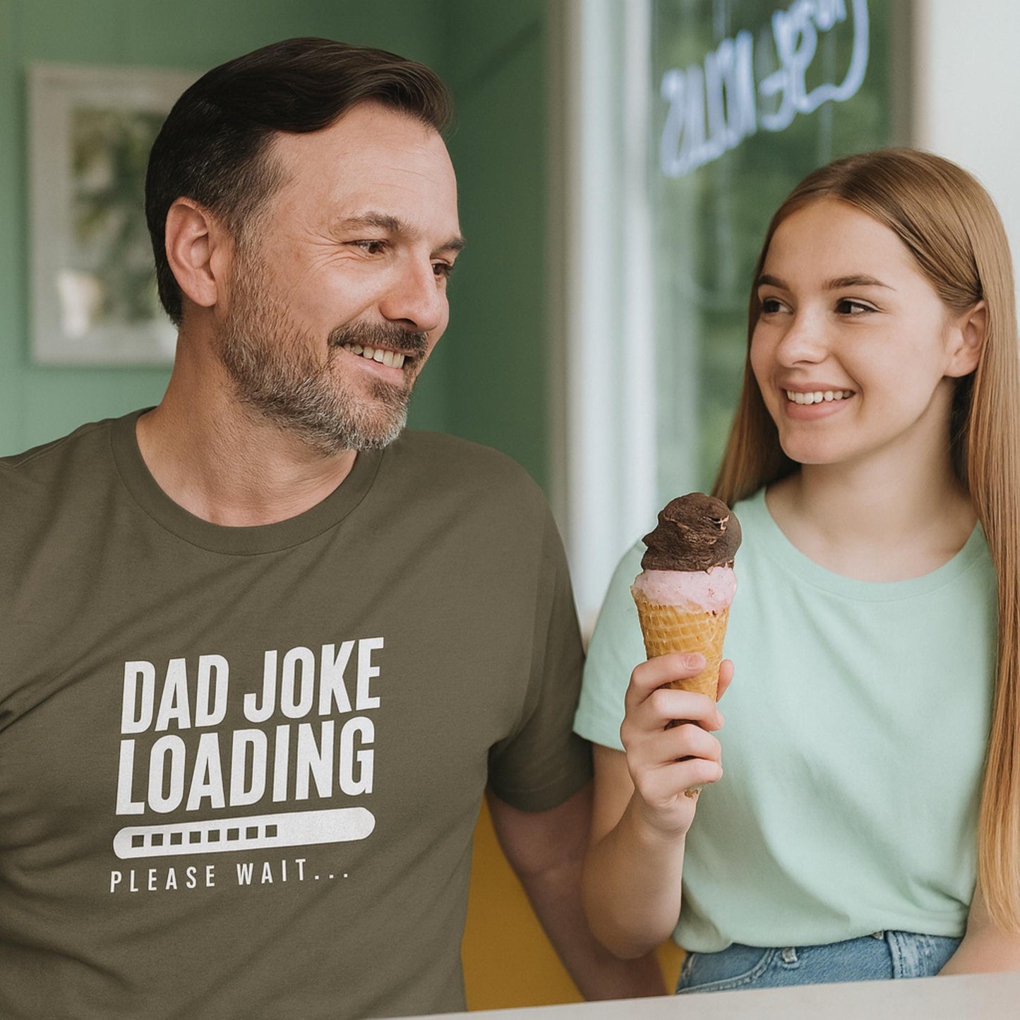 Smiling man wearing an olive green T-shirt with the text “Dad Joke Loading… Please Wait” and a progress bar graphic in white, sitting next to a young woman holding a double-scoop ice cream cone with chocolate and pink ice cream, both looking at each other warmly inside an ice cream shop.