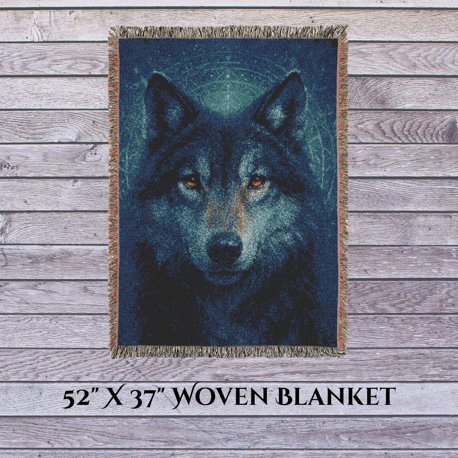 A 52&quot; x 37&quot; woven blanket featuring a striking, realistic image of a wolf’s face with glowing amber eyes. The blanket is displayed vertically against a weathered wood plank background, with multicolored fringe along the edges. Text at the bottom reads “52&#39;&#39; X 37&#39;&#39; WOVEN BLANKET.”