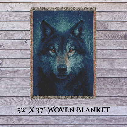 A 52&quot; x 37&quot; woven blanket featuring a striking, realistic image of a wolf’s face with glowing amber eyes. The blanket is displayed vertically against a weathered wood plank background, with multicolored fringe along the edges. Text at the bottom reads “52&#39;&#39; X 37&#39;&#39; WOVEN BLANKET.”