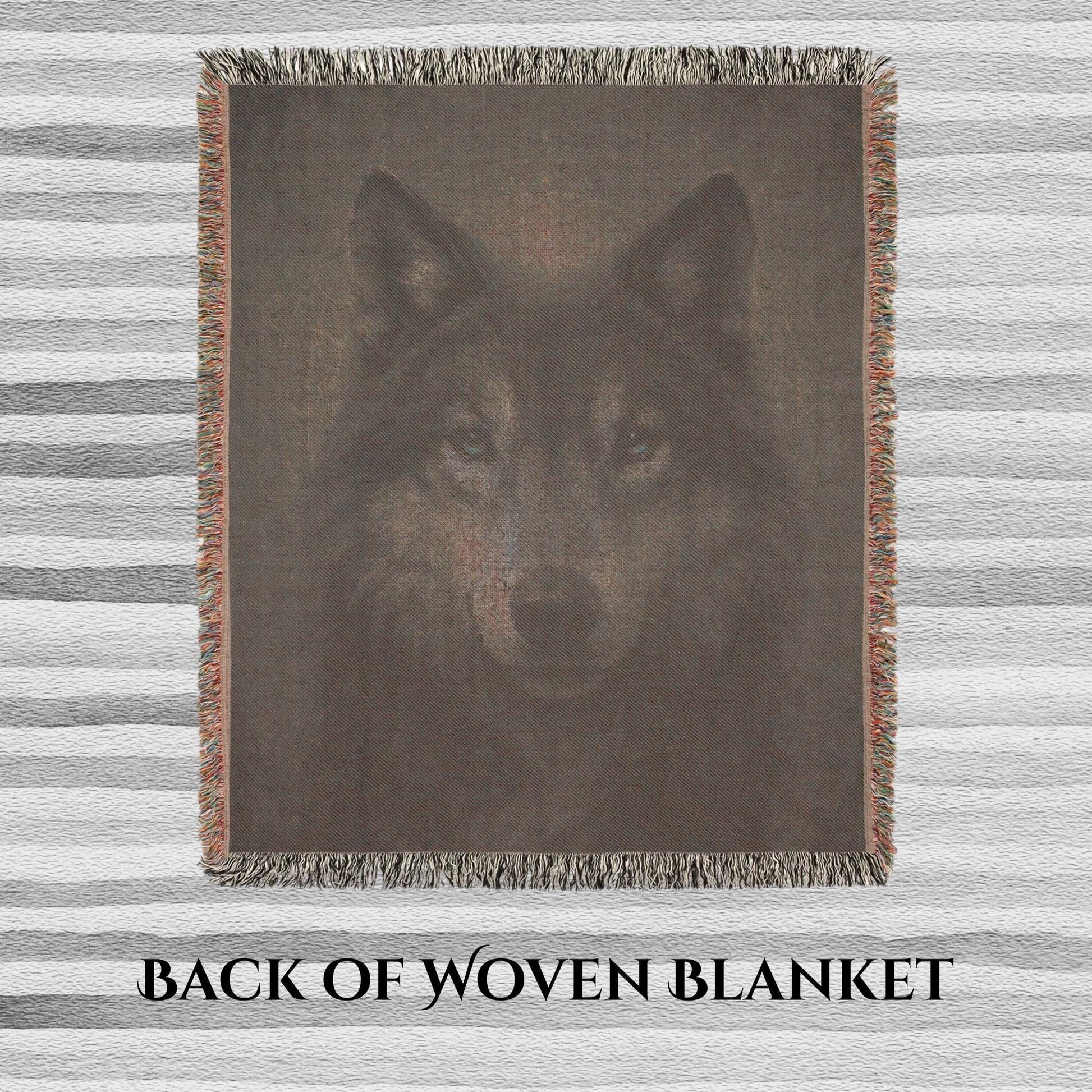 Back side of a woven blanket featuring a muted, reversed image of a wolf&#39;s face with visible thread texture. The blanket has multicolored fringe edges and is laid flat on a striped surface. Text at the bottom reads “BACK OF WOVEN BLANKET.”