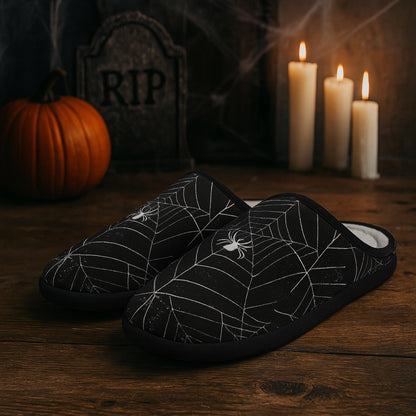Black Halloween-themed slippers with white spiderweb and spider designs sit on a wooden surface. In the background, a tombstone marked &quot;RIP,&quot; a pumpkin, lit candles, and cobwebs create a spooky, festive atmosphere.