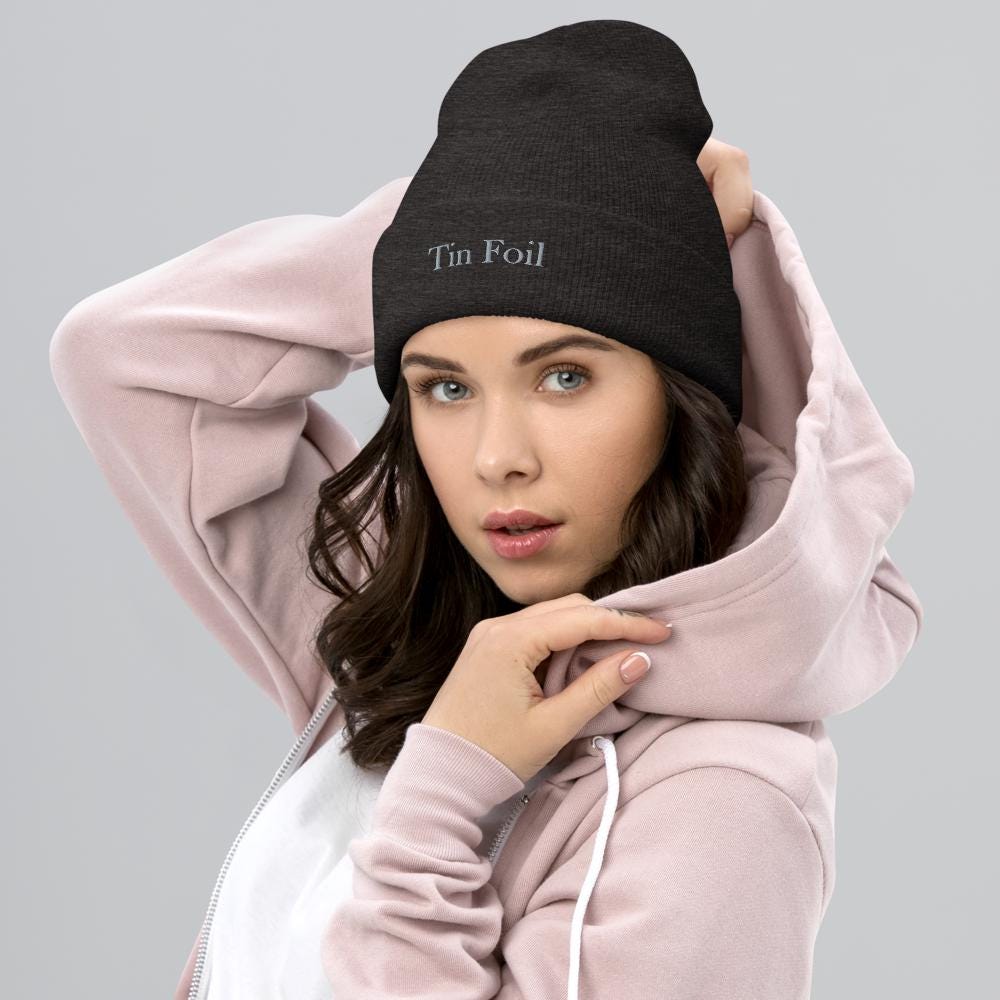 &quot;Woman wearing a black beanie with the text &#39;Tin Foil&#39; embroidered on the front. She has dark hair, light eyes, and is dressed in a light pink hoodie with a white shirt underneath, posing with one hand on her hoodie and the other on her head against a plain grey background.&quot;