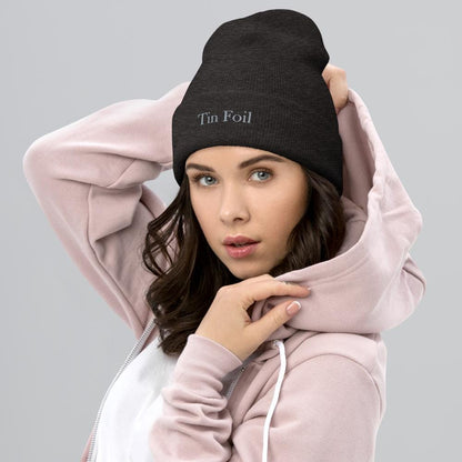 &quot;Woman wearing a black beanie with the text &#39;Tin Foil&#39; embroidered on the front. She has dark hair, light eyes, and is dressed in a light pink hoodie with a white shirt underneath, posing with one hand on her hoodie and the other on her head against a plain grey background.&quot;