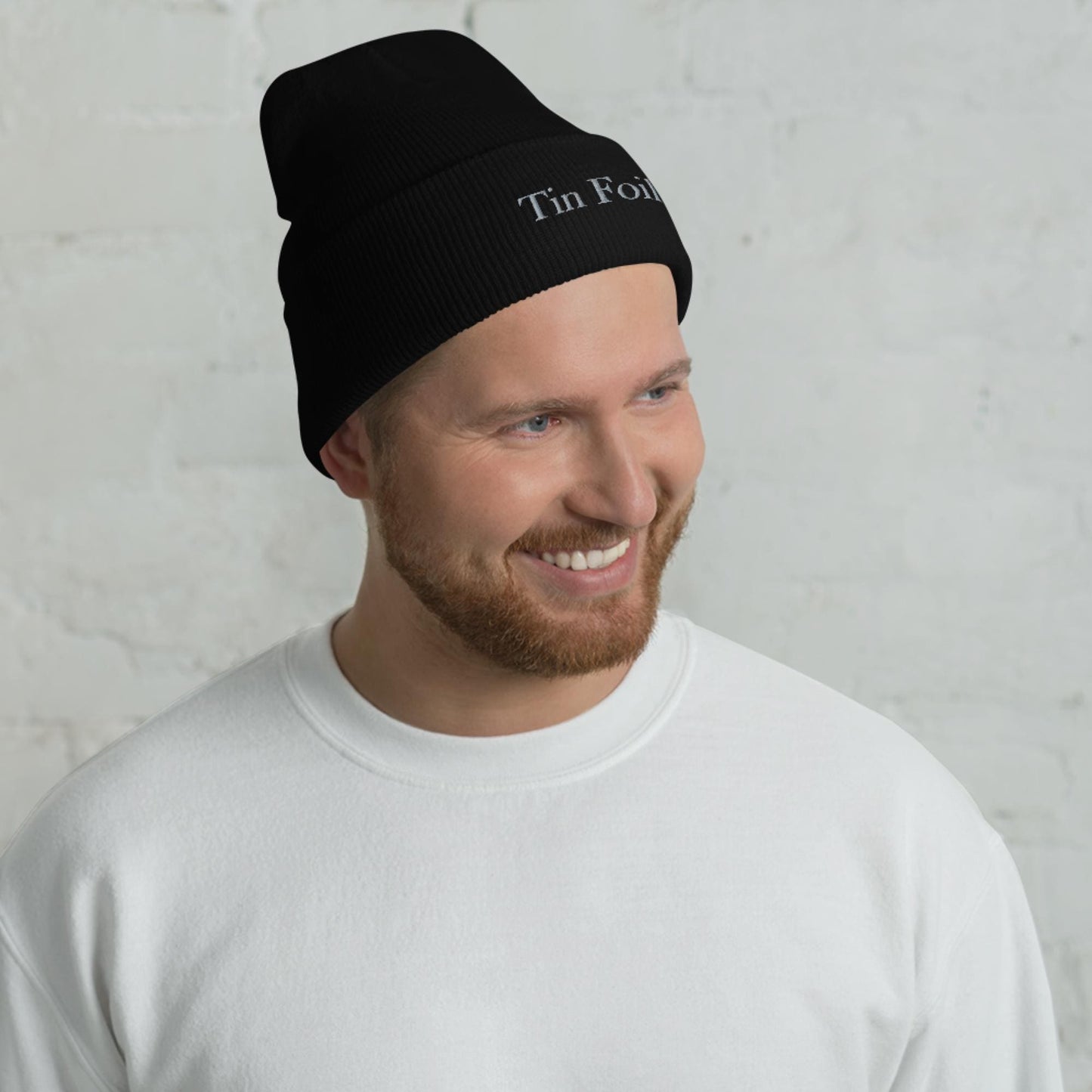 &quot;Smiling man wearing a black beanie with the words &#39;Tin Foil&#39; embroidered on the front. He has a reddish-brown beard and is dressed in a plain white sweatshirt, standing in front of a white brick wall background.&quot;