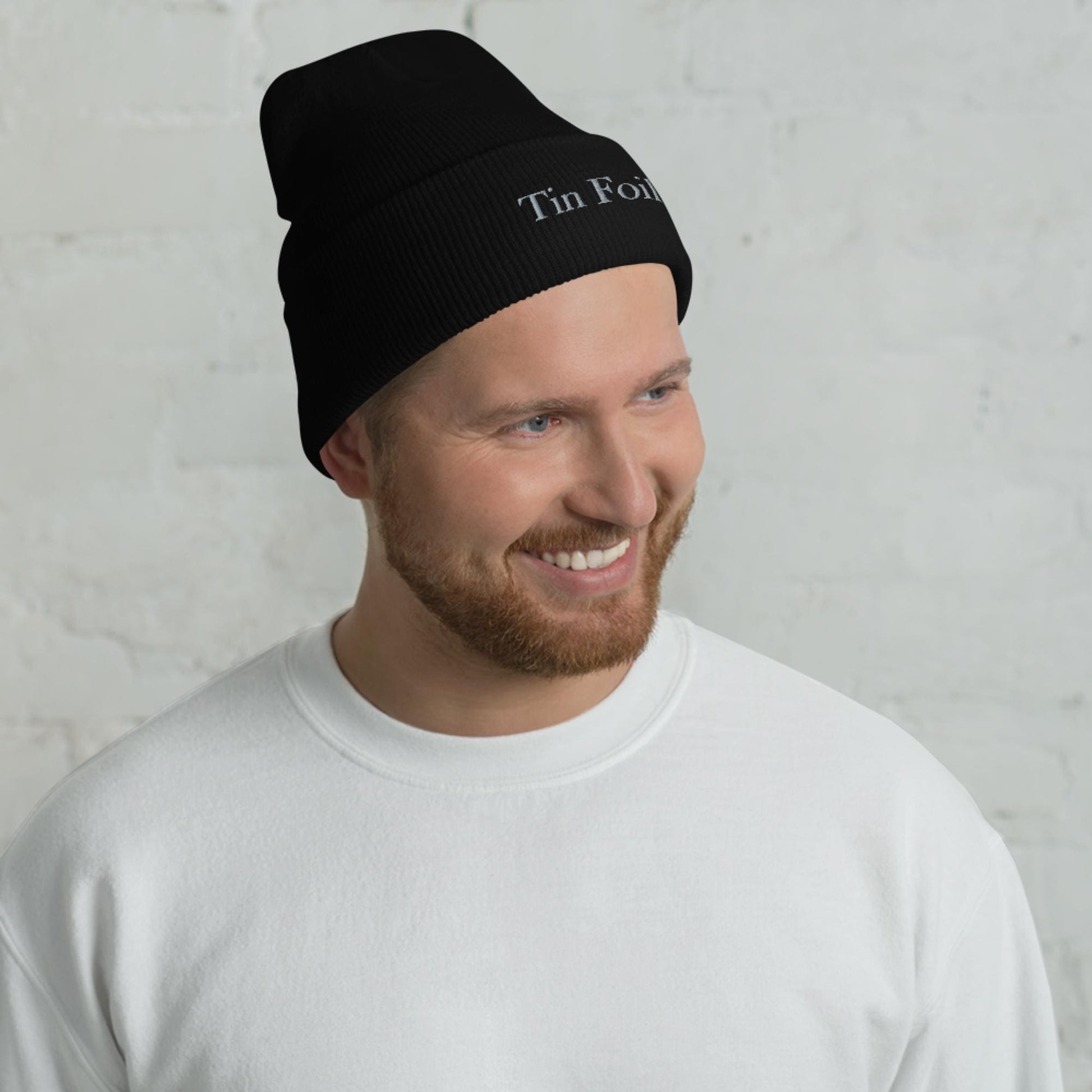 &quot;Smiling man wearing a black beanie with the words &#39;Tin Foil&#39; embroidered on the front. He has a reddish-brown beard and is dressed in a plain white sweatshirt, standing in front of a white brick wall background.&quot;