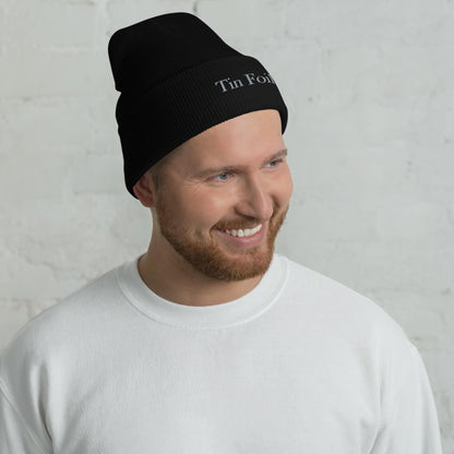 &quot;Smiling man wearing a black beanie with the words &#39;Tin Foil&#39; embroidered on the front. He has a reddish-brown beard and is dressed in a plain white sweatshirt, standing in front of a white brick wall background.&quot;