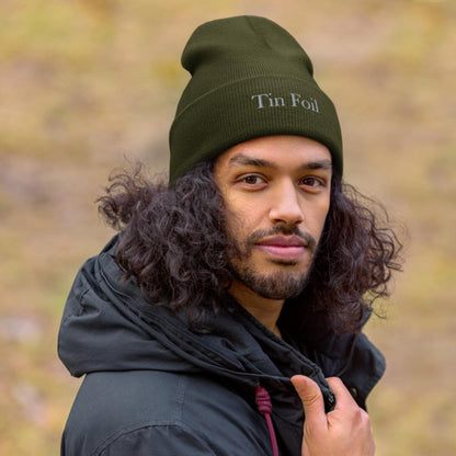 &quot;Man wearing an olive green beanie with the words &#39;Tin Foil&#39; embroidered on the front. He has long curly hair, a trimmed beard, and is dressed in a black hooded jacket, standing outdoors with a soft, blurred natural background.&quot;