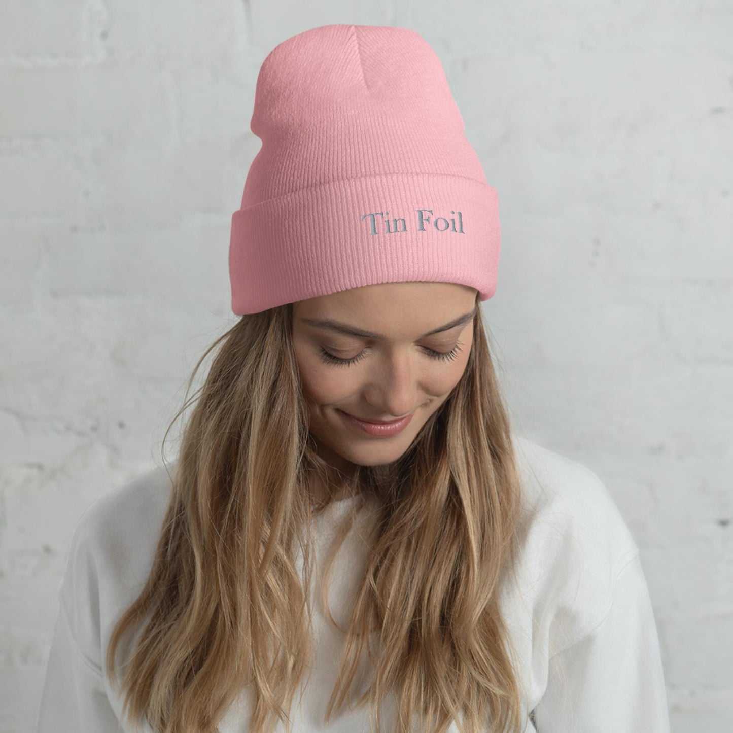 &quot;Woman wearing a pink beanie with the words &#39;Tin Foil&#39; embroidered on the front. She has long blonde hair, is smiling softly while looking down, and is dressed in a white sweatshirt, standing in front of a white brick wall.&quot;