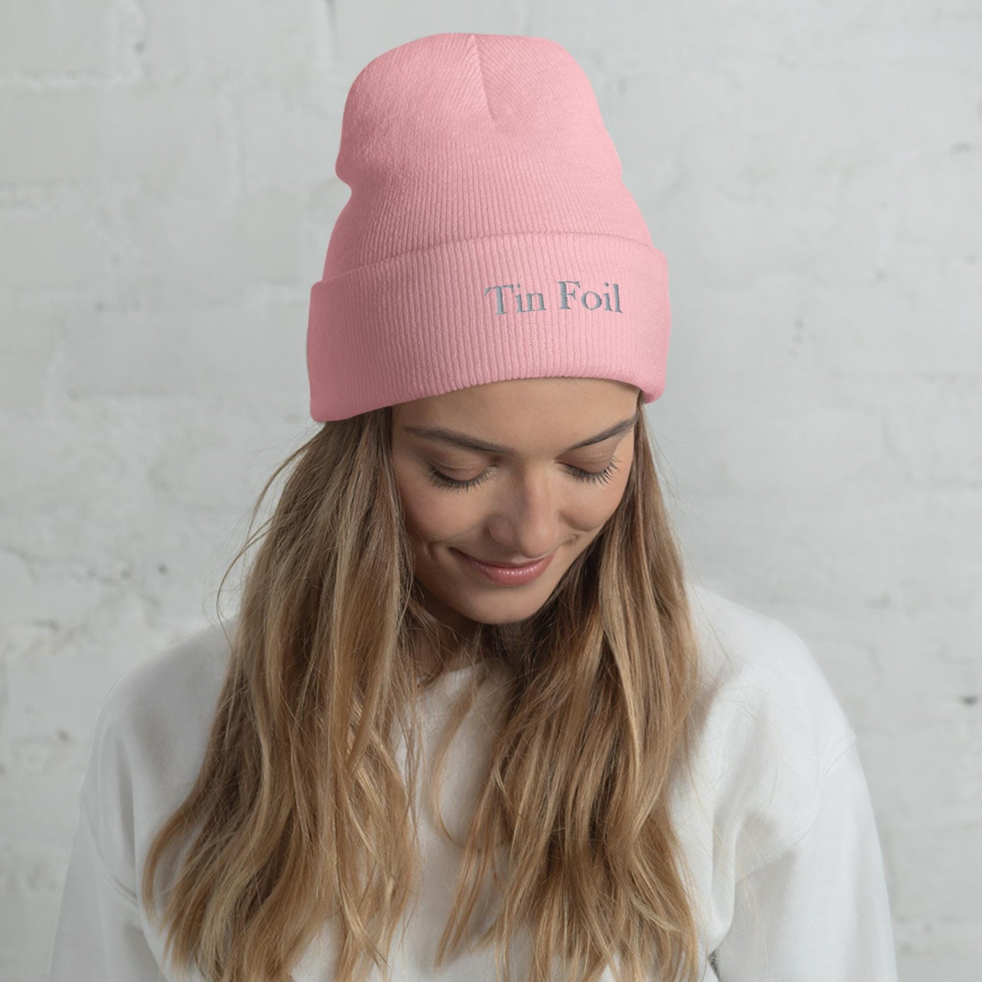 &quot;Woman wearing a pink beanie with the words &#39;Tin Foil&#39; embroidered on the front. She has long blonde hair, is smiling softly while looking down, and is dressed in a white sweatshirt, standing in front of a white brick wall.&quot;