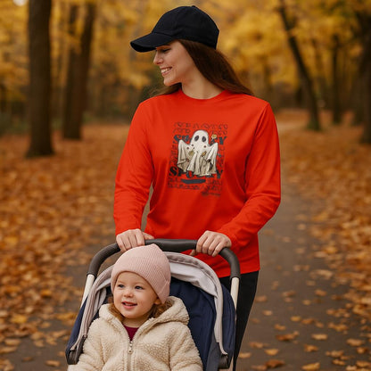 Woman smiling while pushing a stroller along a tree-lined path covered in autumn leaves. She is wearing a navy baseball cap and an orange long-sleeve shirt featuring a ghost graphic surrounded by candy with the repeated words &#39;SEASON&#39; and &#39;SPOOKY.&#39; A young child in the stroller wears a pink knit beanie and a fuzzy cream-colored jacket, smiling and enjoying the fall day.