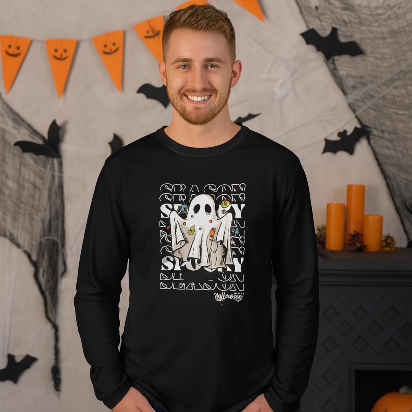 Smiling man wearing a black long-sleeve shirt with a Halloween-themed design featuring a cartoon ghost surrounded by candy, set against a repeating &#39;SEASON&#39; and &#39;SPOOKY&#39; text background. He is standing in a festive room decorated with orange candles, black bats, cobwebs, and a triangular orange banner with smiley faces.