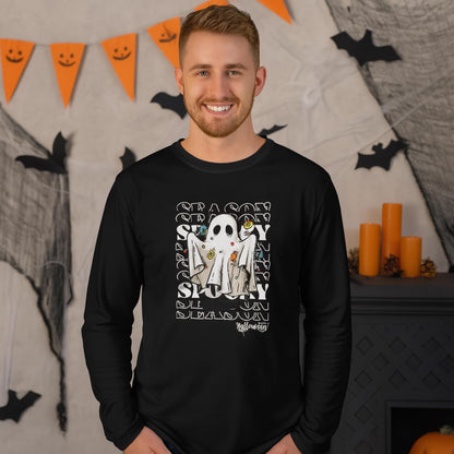 Smiling man wearing a black long-sleeve shirt with a Halloween-themed design featuring a cartoon ghost surrounded by candy, set against a repeating &#39;SEASON&#39; and &#39;SPOOKY&#39; text background. He is standing in a festive room decorated with orange candles, black bats, cobwebs, and a triangular orange banner with smiley faces.
