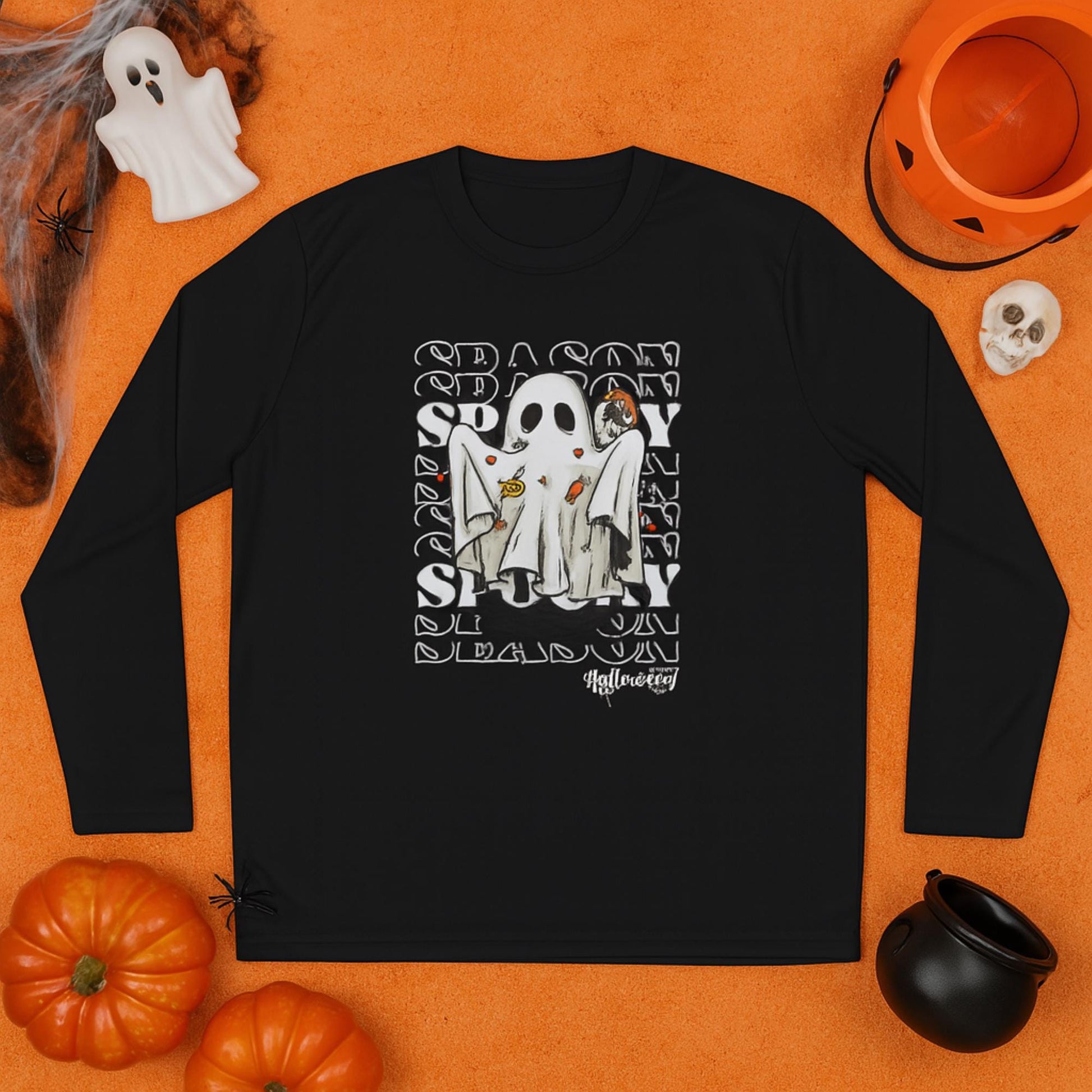 Black long-sleeve shirt laid flat on an orange surface, featuring a Halloween-themed design of a ghost costume with candy, set against a repeating &#39;SEASON SPOOKY&#39; text background. Surrounding the shirt are festive decorations including pumpkins, a jack-o&#39;-lantern bucket, a small skull, a ghost figurine, fake cobwebs with a spider, and a black cauldron.