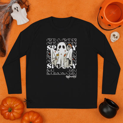 Black long-sleeve shirt laid flat on an orange surface, featuring a Halloween-themed design of a ghost costume with candy, set against a repeating &#39;SEASON SPOOKY&#39; text background. Surrounding the shirt are festive decorations including pumpkins, a jack-o&#39;-lantern bucket, a small skull, a ghost figurine, fake cobwebs with a spider, and a black cauldron.