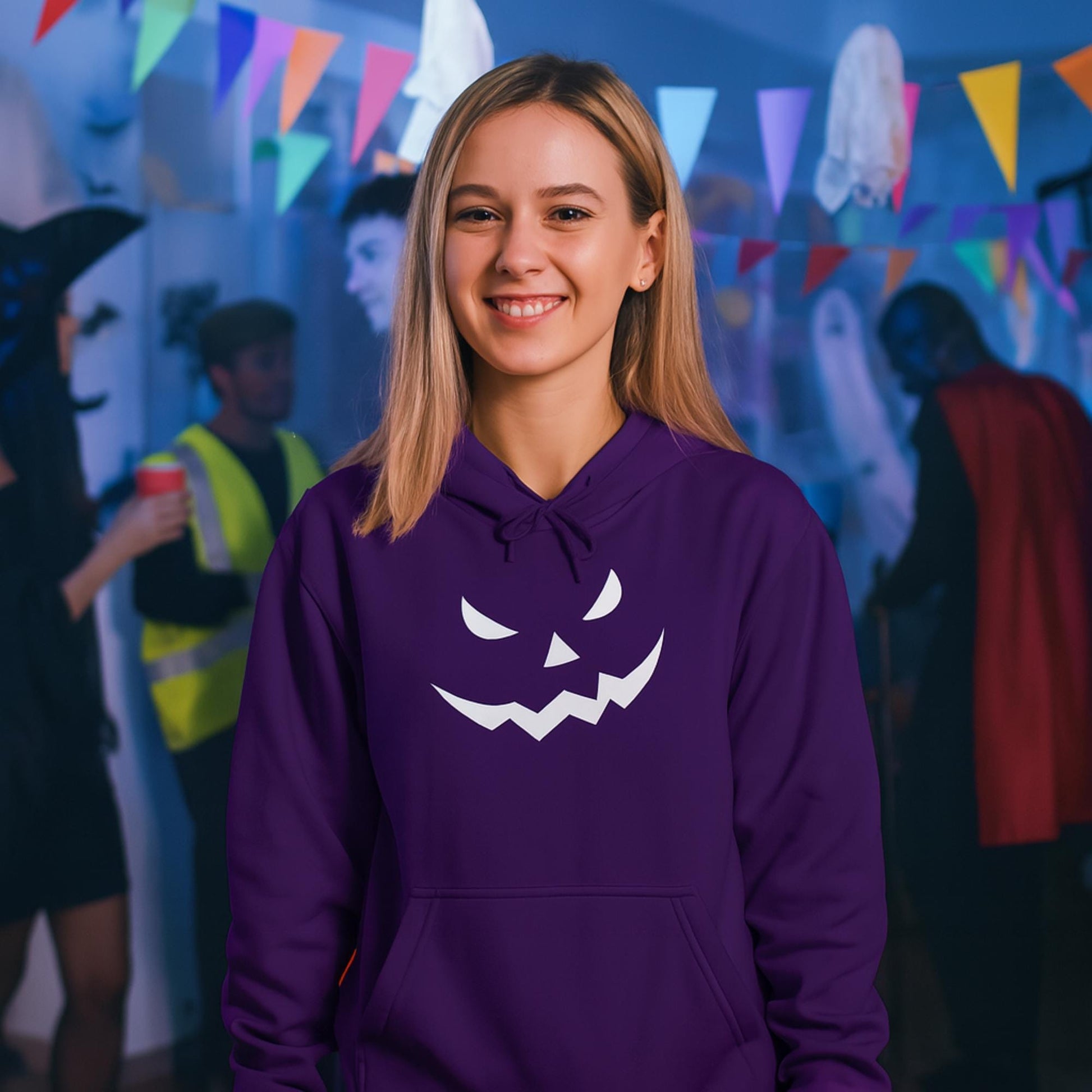 Smiling woman wearing a purple hoodie with a white jack-o&#39;-lantern face design on the front, standing at a Halloween party. The background features partygoers in costumes, colorful triangular banners, and festive decorations including ghost cutouts and lights.