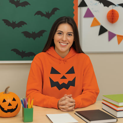 Smiling woman sitting at a classroom desk wearing an orange hoodie with a black jack-o&#39;-lantern face design on the front. The classroom is decorated with Halloween-themed items including a green chalkboard with black bat cutouts, a whiteboard with colorful triangular bunting, and a carved pumpkin on the desk. Notebooks, pencils, and a stack of books are also on the desk.