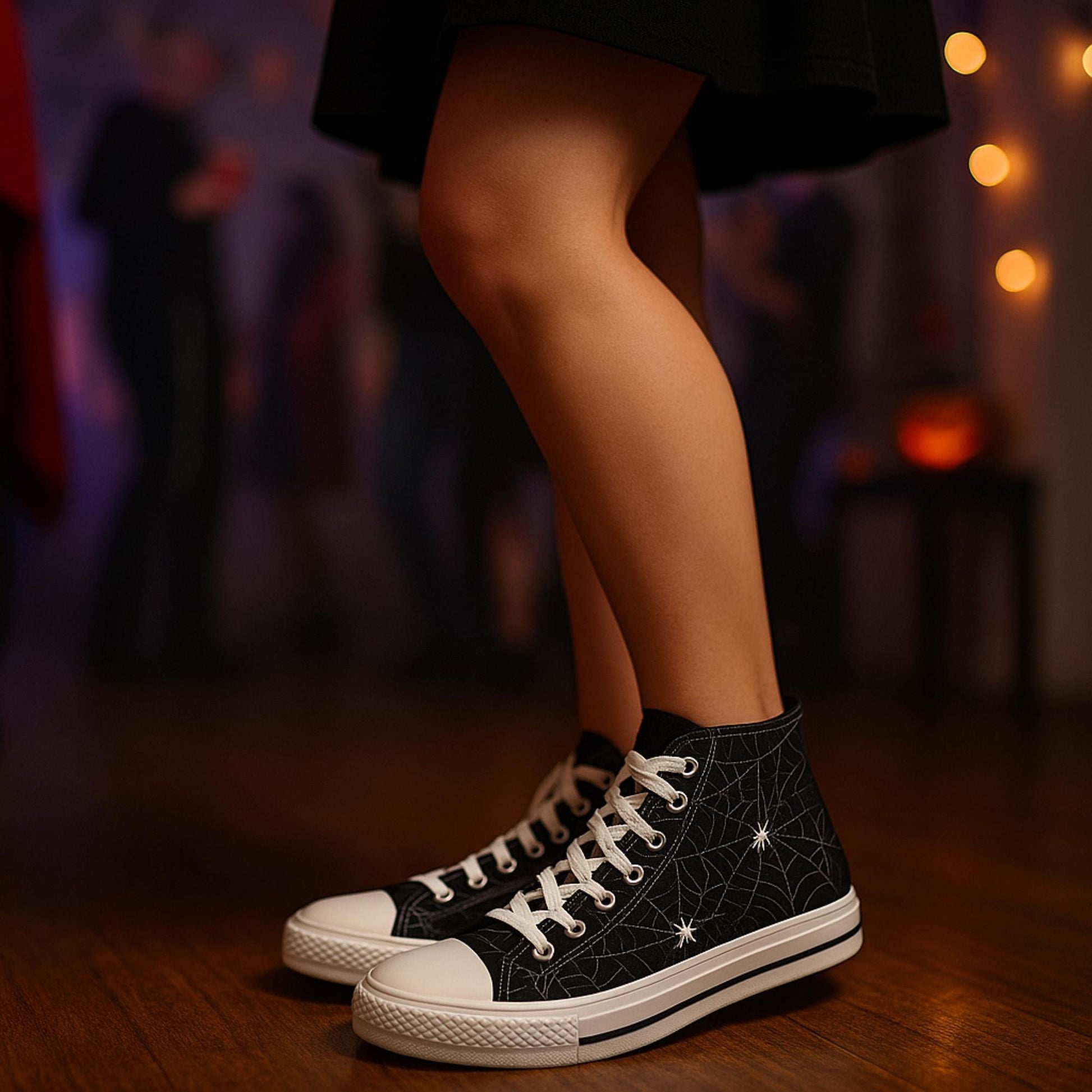 Close-up of a person wearing black high-top sneakers with a white spiderweb and spider design, standing indoors on a wooden floor. The person is dressed in a black skirt, and the background is softly blurred, showing people at a Halloween party with dim lighting and festive decorations.