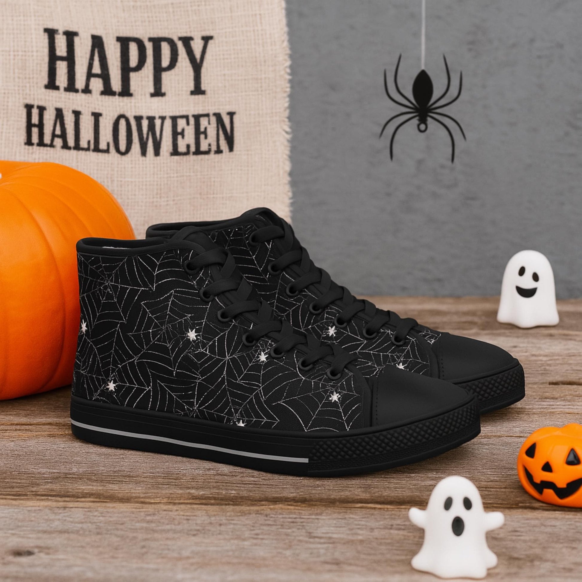 Pair of black high-top sneakers with black soles, toe caps, and laces, featuring a white spiderweb and spider pattern. The shoes are displayed on a wooden surface with Halloween decorations including pumpkins, ghost figures, a hanging spider, and a burlap sign that reads &#39;Happy Halloween&#39; in the background.