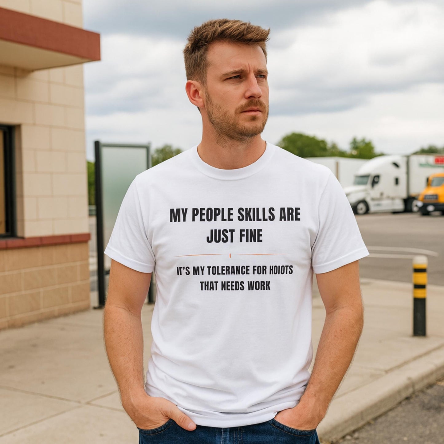 Man wearing a white T-shirt with bold black and orange text that reads, “MY PEOPLE SKILLS ARE JUST FINE — IT’S MY TOLERANCE FOR IDIOTS THAT NEEDS WORK.” He stands outdoors in front of a building and a parking lot with semi-trucks in the background, looking off into the distance with a serious expression.