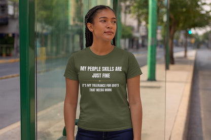 Woman standing at a bus stop wearing an olive green T-shirt that reads, “MY PEOPLE SKILLS ARE JUST FINE — IT’S MY TOLERANCE FOR IDIOTS THAT NEEDS WORK” in bold white text. She looks off into the distance with a neutral expression, surrounded by a quiet urban street scene with glass panels and green poles.