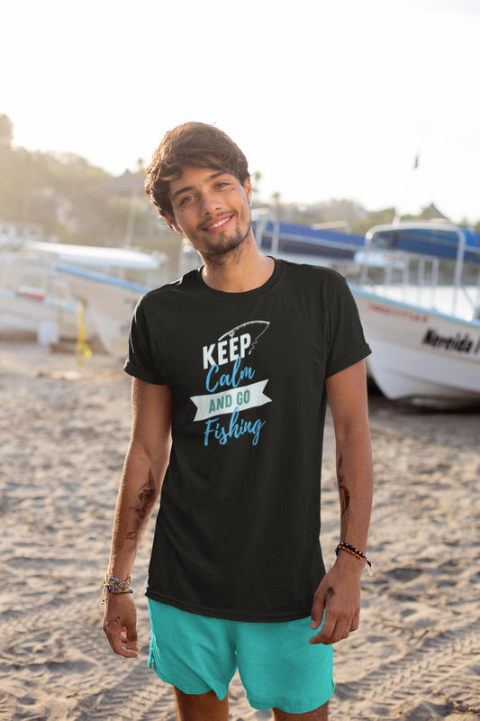 Young man standing on a sandy beach near boats, wearing a black T-shirt with a playful fishing-themed design. The shirt features the text “KEEP Calm AND GO Fishing” in white, teal, and blue fonts, with a fishing hook graphic above the word “KEEP.” He is also wearing teal shorts and smiling at the camera in the warm, coastal sunset light.