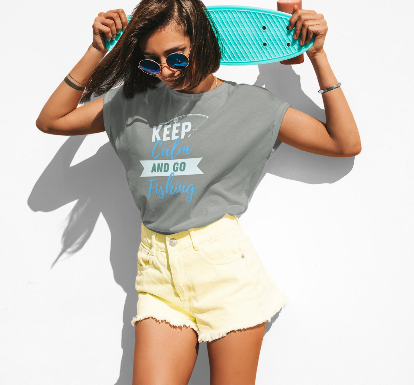 Stylish young woman in sunglasses holding a turquoise skateboard behind her neck, wearing a relaxed gray T-shirt with the phrase “KEEP Calm AND GO Fishing” in white, teal, and blue text along with a fishing hook graphic. She pairs the shirt with high-waisted pale yellow denim shorts and poses confidently against a bright white background.