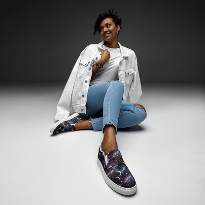 Stylish woman sitting on the floor wearing galaxy-themed slip-on shoes with white soles. The shoes feature a cosmic design with stars, nebulae, and crescent moons in shades of purple, blue, and black. She is dressed in light blue ripped jeans, a white t-shirt, and a white denim jacket, striking a relaxed and confident pose against a minimalist gray background.