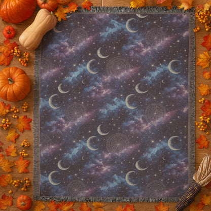 Flat-lay of a celestial-themed woven blanket with a design featuring crescent moons, stars, and nebula clouds in shades of purple, blue, and black. The blanket is bordered with fringe and surrounded by fall-themed decorations including pumpkins, autumn leaves, berries, and a gourd, creating a cozy seasonal aesthetic.