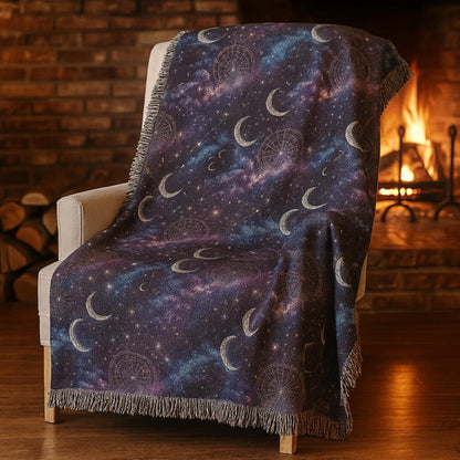 A cozy woven blanket with a celestial design featuring crescent moons, stars, and constellation patterns in a galaxy-inspired palette of deep purples, blues, and blacks. The blanket is draped over a cream armchair in front of a lit fireplace, with a rustic brick wall and stacked firewood in the background, creating a warm and inviting atmosphere.