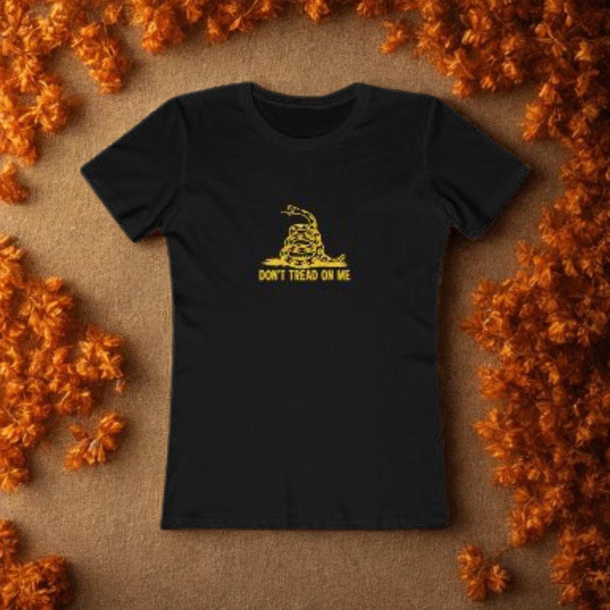 Black fitted t-shirt laid flat on a brown surface, surrounded by autumn orange leaves. The shirt features a yellow graphic of a coiled rattlesnake and the text &quot;DON&#39;T TREAD ON ME&quot; in capital letters, referencing the Gadsden flag.