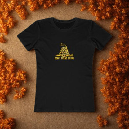 Black fitted t-shirt laid flat on a brown surface, surrounded by autumn orange leaves. The shirt features a yellow graphic of a coiled rattlesnake and the text &quot;DON&#39;T TREAD ON ME&quot; in capital letters, referencing the Gadsden flag.