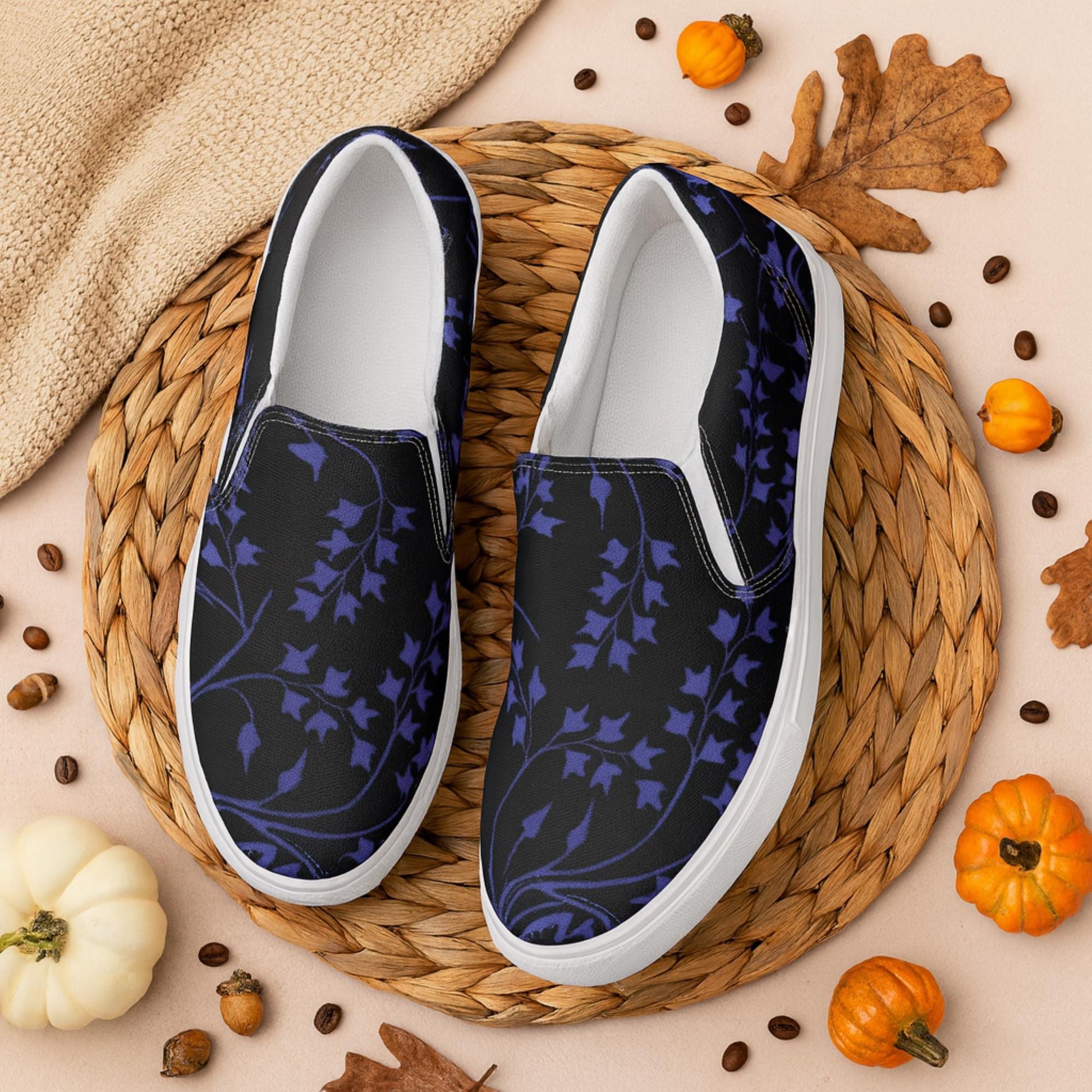 Pair of black slip-on shoes with a dark purple ivy vine pattern, displayed on a round woven placemat surrounded by small pumpkins, autumn leaves, coffee beans, and cozy fall decor. The setting evokes a warm, seasonal autumn atmosphere.