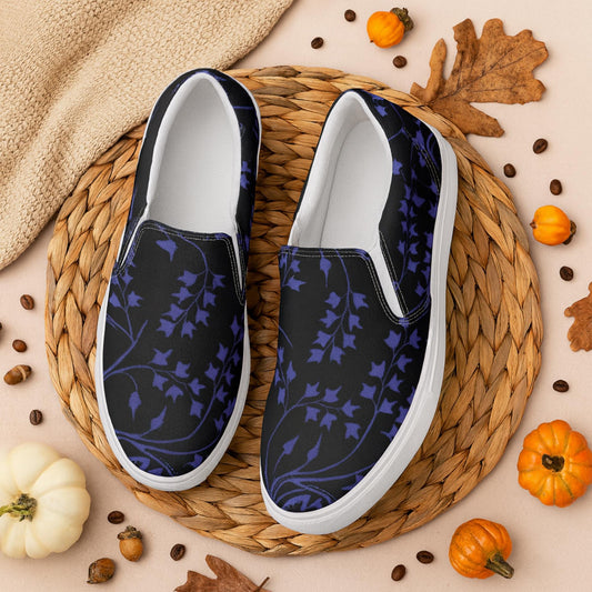 Pair of black slip-on shoes with a dark purple ivy vine pattern, displayed on a round woven placemat surrounded by small pumpkins, autumn leaves, coffee beans, and cozy fall decor. The setting evokes a warm, seasonal autumn atmosphere.