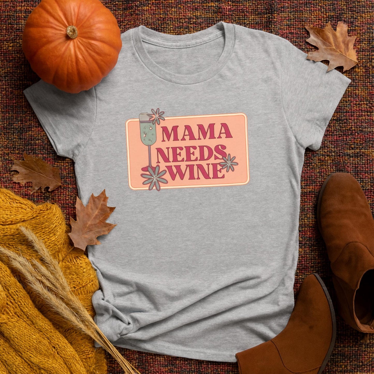 Flat lay of a heather gray women&#39;s t-shirt with a retro-style graphic that reads &quot;Mama Needs Wine&quot; in bold pink text, featuring a champagne glass and whimsical flowers on a peach background. The shirt is styled on a fall-themed woven blanket with seasonal props including a small pumpkin, brown leather boots, dried wheat, orange knitwear, and scattered autumn leaves.