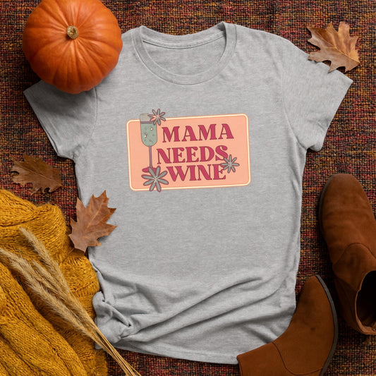 Flat lay of a heather gray women&#39;s t-shirt with a retro-style graphic that reads &quot;Mama Needs Wine&quot; in bold pink text, featuring a champagne glass and whimsical flowers on a peach background. The shirt is styled on a fall-themed woven blanket with seasonal props including a small pumpkin, brown leather boots, dried wheat, orange knitwear, and scattered autumn leaves.